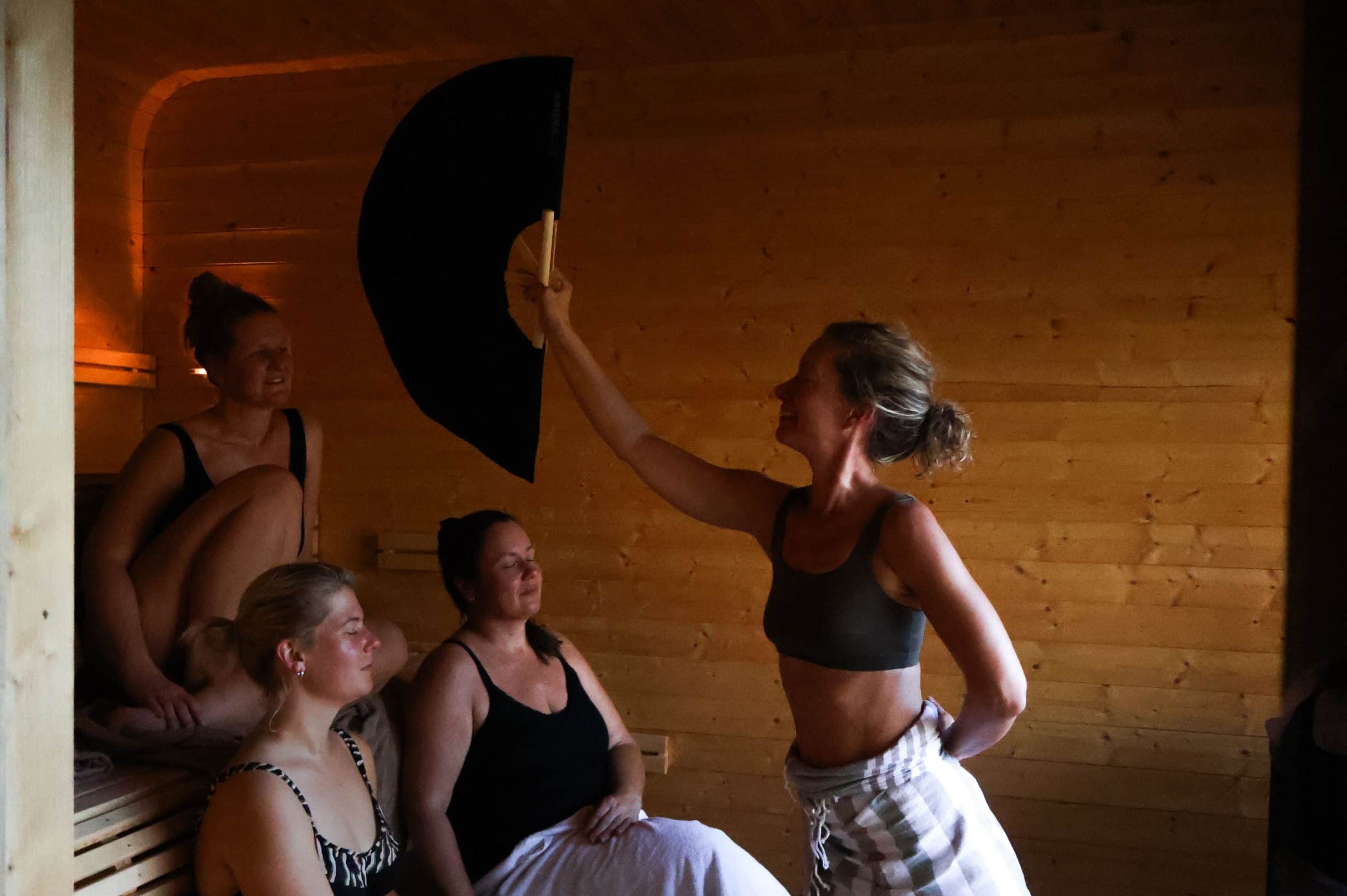 Kvinne som vifter damp fra badstueovn mot to kvinner som sitter i badstue