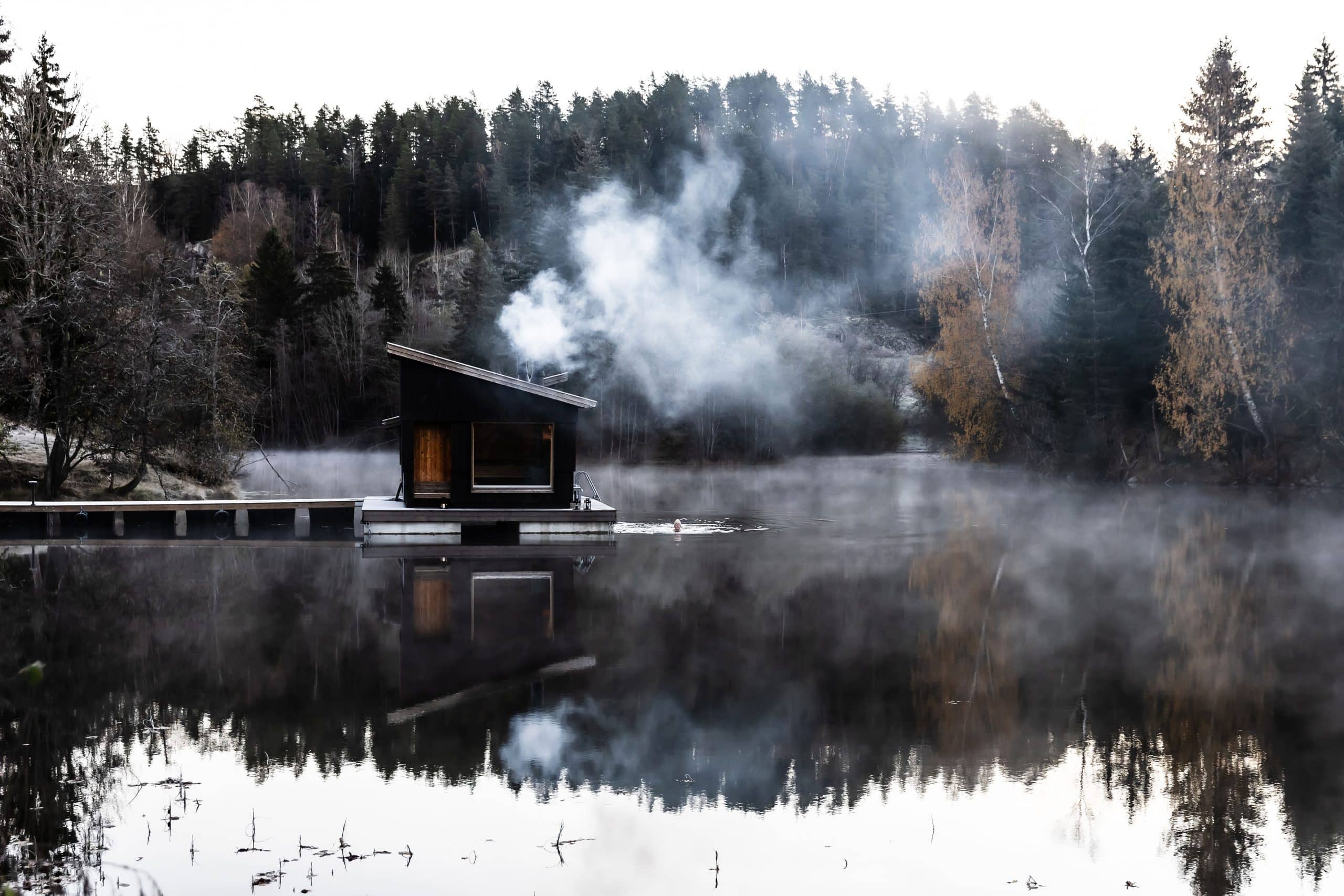 Flytende badstue med røyk fra pipa en kald høstmorgen. Noen svømmer i vannet med lue på.